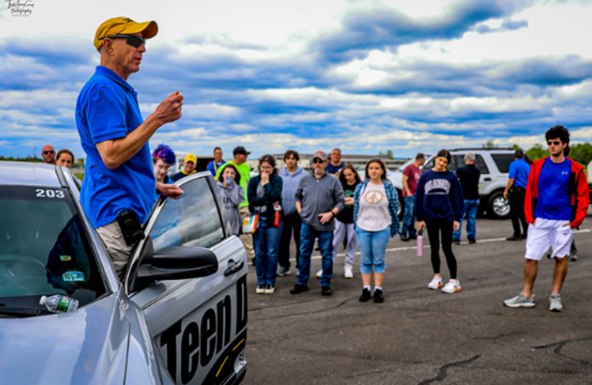 Person next to a car speaking to a group of youth standing in a circle around him