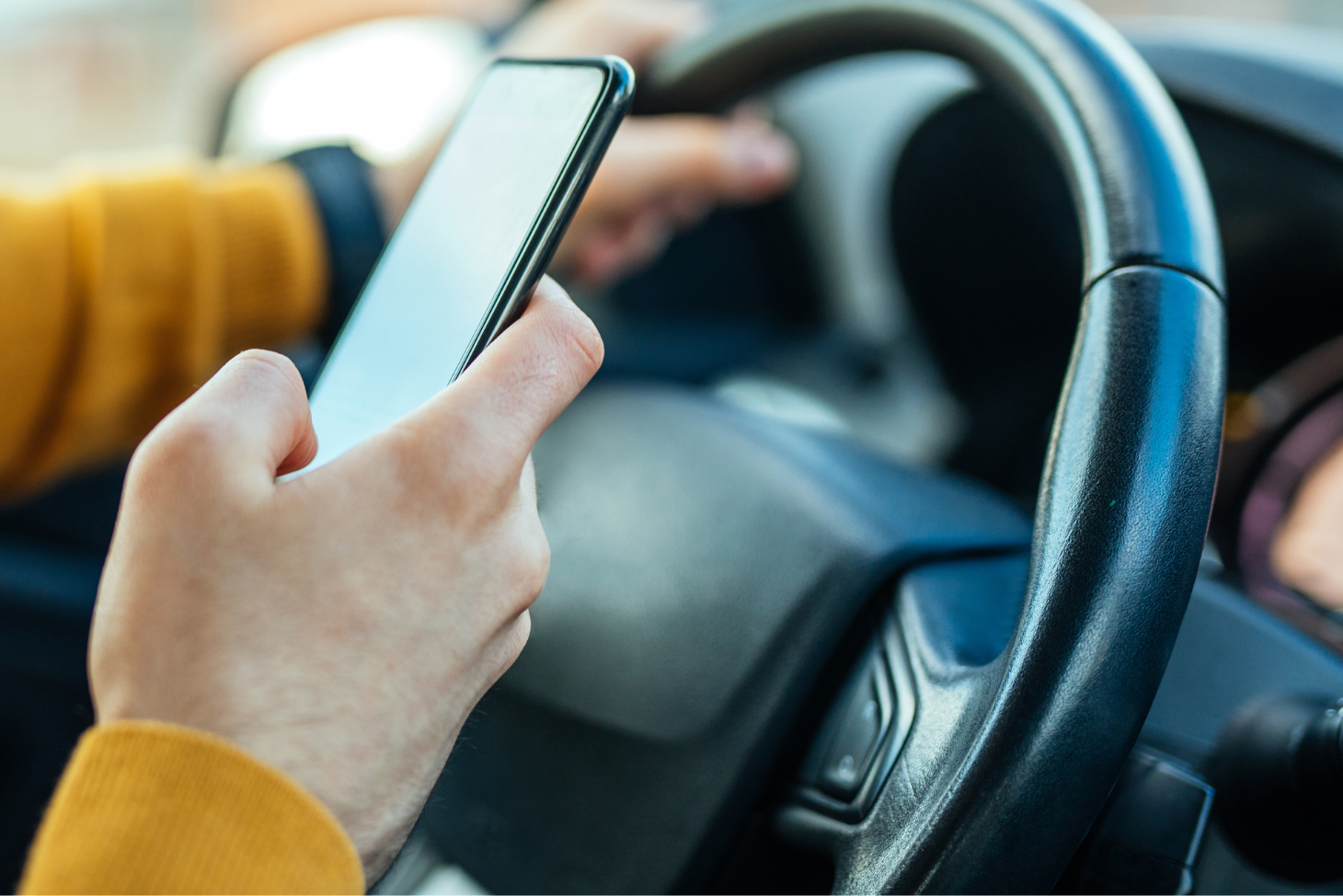 A hand holds a cell phone in front of a steering wheel