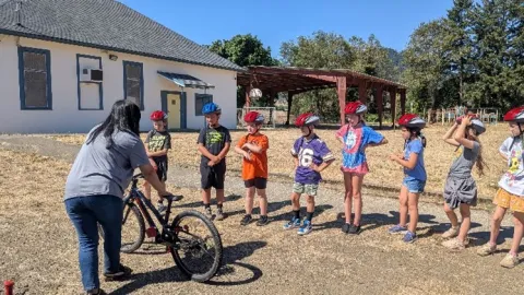 Kids wearing helmets in a line receiving safe bike riding instructions from an adult